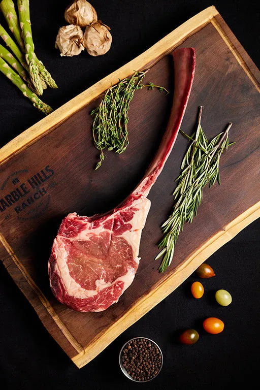 A USDA Tomahawk Ribeye steak displayed on a cutting board, accompanied by herbs and vegetables.