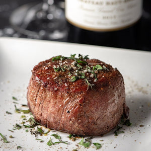 A Filet Mignon steak on a white plate with herbs and a bottle of wine.